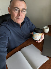 A man in a navy quarter-zip holds a jar of CappyRoots Focus gummies at a wooden desk beside an open notebook and a mug