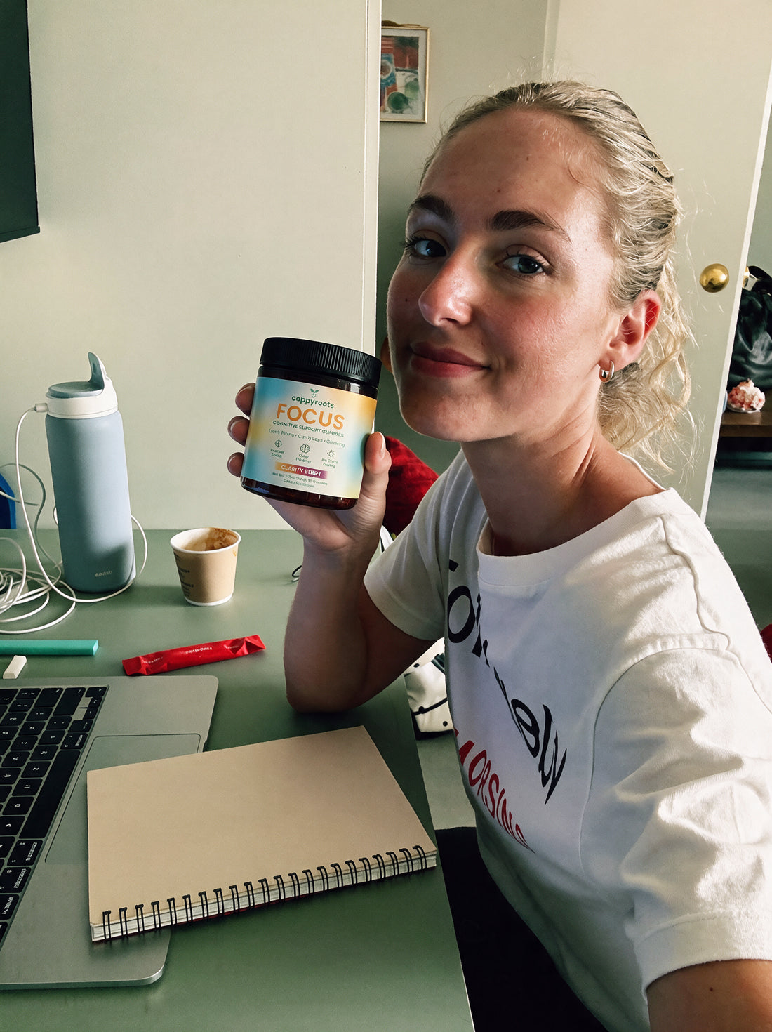 A woman at a home desk holds up a jar of CappyRoots Focus gummies beside a notebook and water bottle