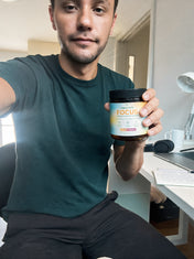 A man in a green tank top holds a jar of CappyRoots Focus gummies beside a desk