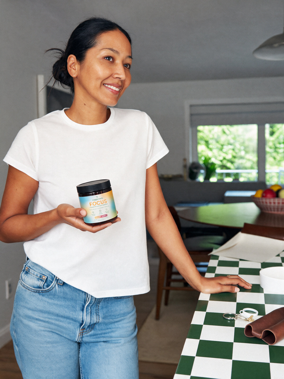 A woman in a white t-shirt and jeans holds a jar of CappyRoots Focus gummies in a kitchen